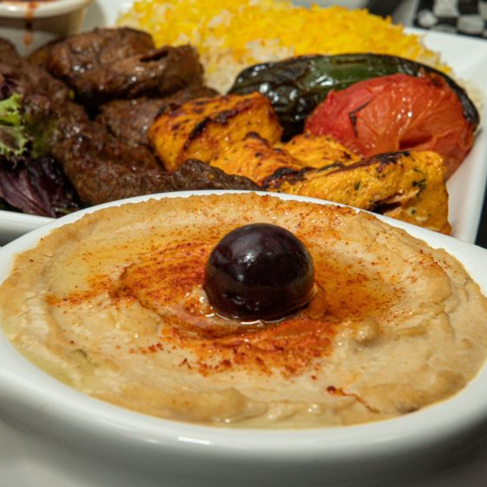 A bowl of hummus with an olive and paprika, with a plate of Mediterranean kebabs in the background.
